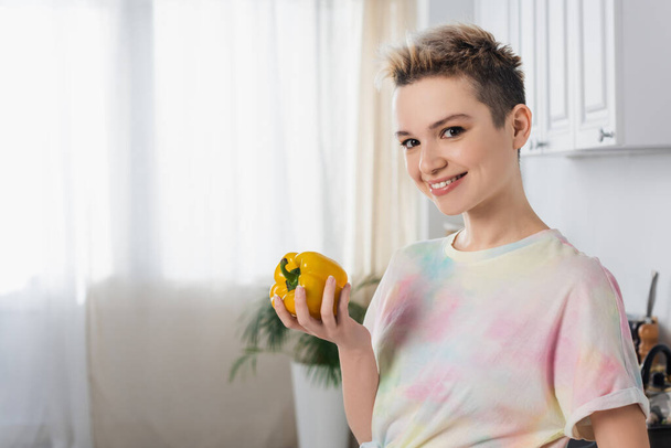happy pangender person holding whole bell pepper and looking at camera in kitchen - Φωτογραφία, εικόνα