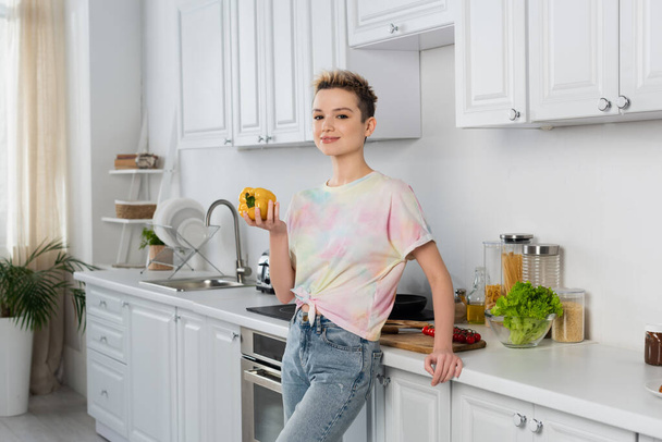 bigender person with short hair smiling at camera while holding whole bell pepper in kitchen - Photo, Image