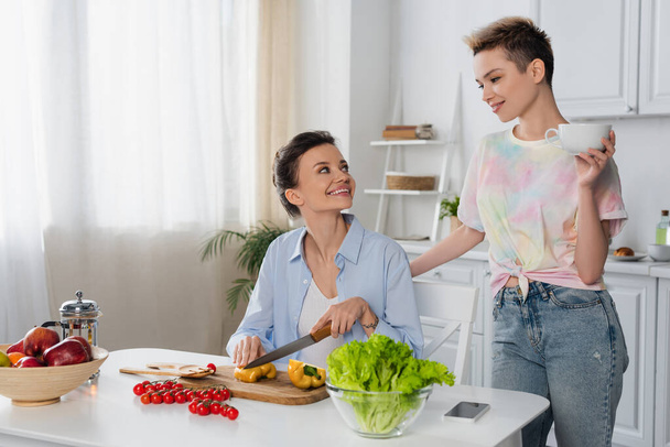 cheerful pangender person cutting fresh vegetables near lover with cup of tea - Photo, Image