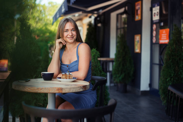 smiling girl at the camera sits in a cafe for a cup of coffee among green cypresses. - Photo, Image