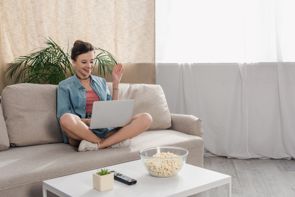 mujer sonriente sentada en el sofá con las piernas cruzadas y la mano agitada durante la videollamada en el ordenador portátil - Foto, imagen