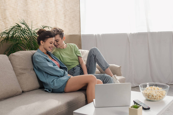 happy pansexual lovers sitting on couch near coffee table with laptop and popcorn - Photo, Image