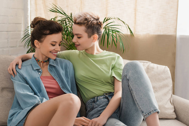 happy pangender lovers embracing and holding hands on couch at home - Фото, зображення
