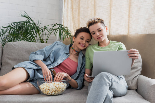 pleased pansexual couple watching film on laptop near bowl of popcorn on couch - Photo, Image