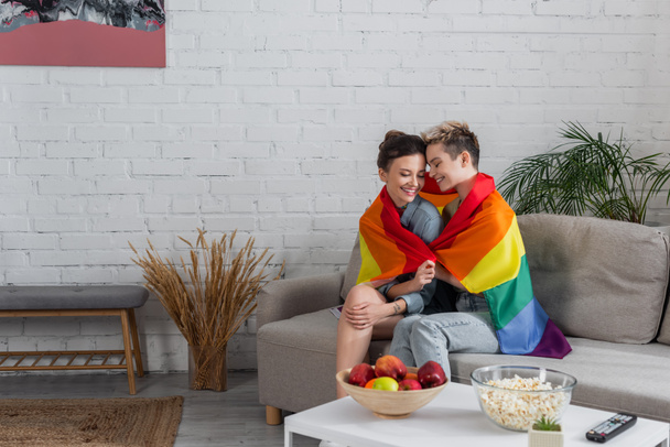 happy pangender couple sitting on couch under lgbt flag near apples and popcorn on coffee table - Foto, Imagen