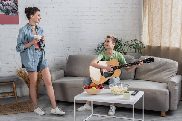 bigender hudebník hraje na kytaru na gauči poblíž usmívající se partner stojící s šálkem čaje - Fotografie, Obrázek