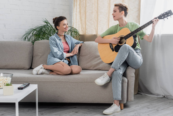 young pansexual person playing guitar near lover on sofa in living room - Foto, Imagem