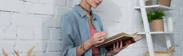 vista parcial de la mujer tatuada en el libro de lectura de camisa de mezclilla cerca de la pared de ladrillo blanco, pancarta - Foto, Imagen