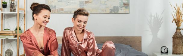θετική pansexual άτομα σε ρόμπα και σατέν πιτζάμες χαμογελώντας στην κρεβατοκάμαρα, πανό - Φωτογραφία, εικόνα