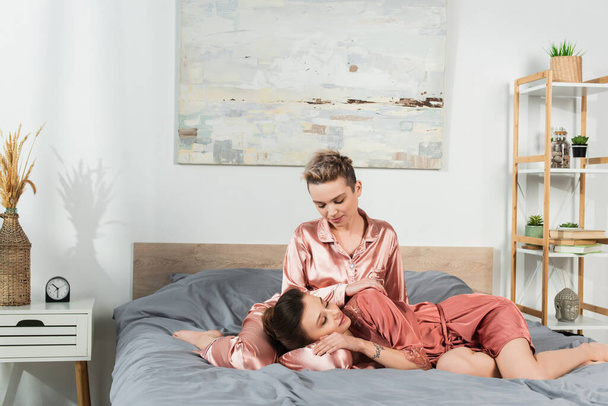 young pangender person resting on bed near partner in satin pajamas - Foto, imagen