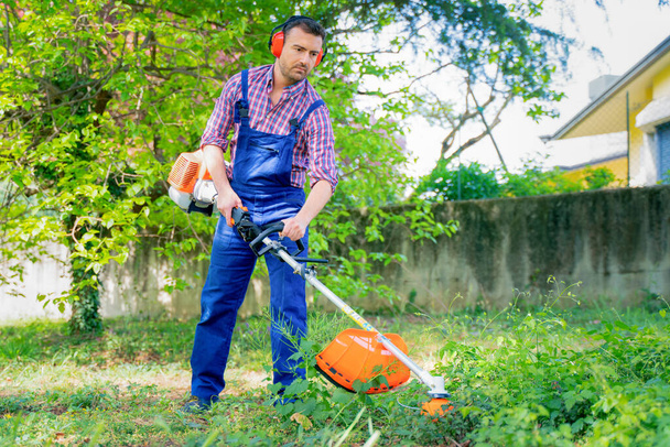 Ένας άνδρας που εργάζεται στον κήπο και κουρεύει γρασίδι χρησιμοποιώντας θαμνοκοπτικό - Φωτογραφία, εικόνα