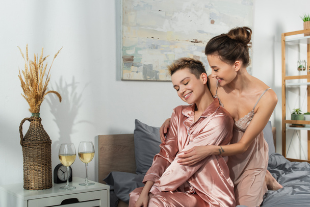 smiling bigender person hugging young lover sitting in silk pajamas near glasses of white wine - Foto, immagini