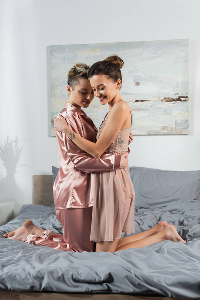 young and happy pangender couple in sleepwear embracing on bed at home - 写真・画像