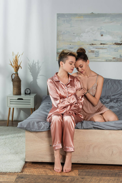 young bigender person leaning on lover sitting on bed in satin pajamas - Valokuva, kuva