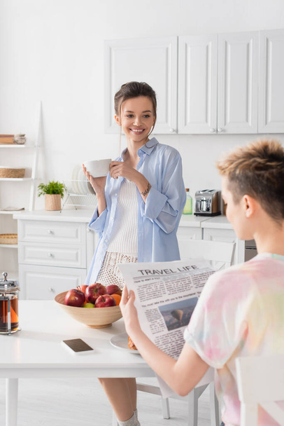 cheerful pansexual person standing with tea cup near blurred partner reading newspaper during breakfast - Foto, Bild