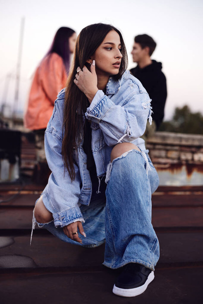 Дівчинка-підліток в джинсовому вбранні присідає на даху будівлі, під час перебування зі своїми друзями
. - Фото, зображення