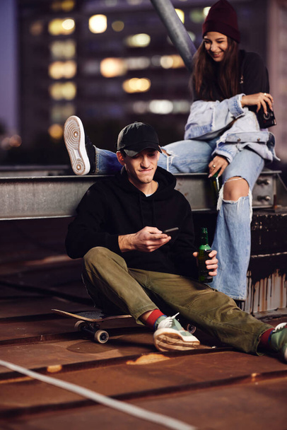 Ένας έφηβος σκέιτερ αγόρι και η φίλη του κάθονται στην οροφή του κτιρίου πίνοντας μπύρα και διασκεδάζοντας τη νύχτα έξω. - Φωτογραφία, εικόνα