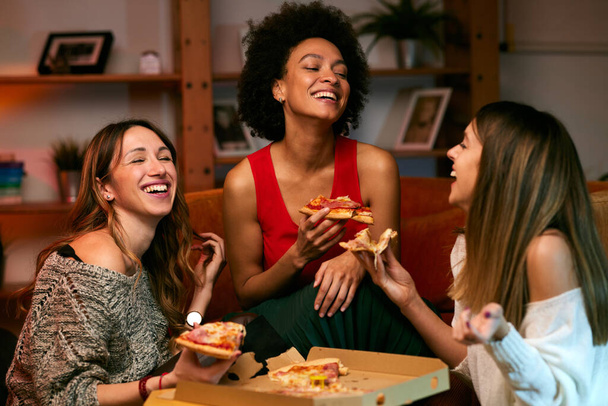 Monikulttuurinen ryhmä iloisia nuoria naisia syö pizzaa istuessaan olohuoneessa.. - Valokuva, kuva
