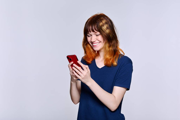 Smiling teenage female looking at smartphone screen, on light studio background - Foto, imagen