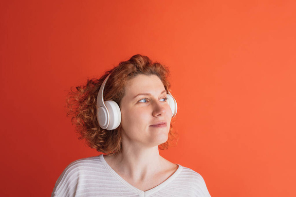 Portrait of calmly looking red-haired woman listening to music in white headphone isolated over red studio background - Foto, immagini