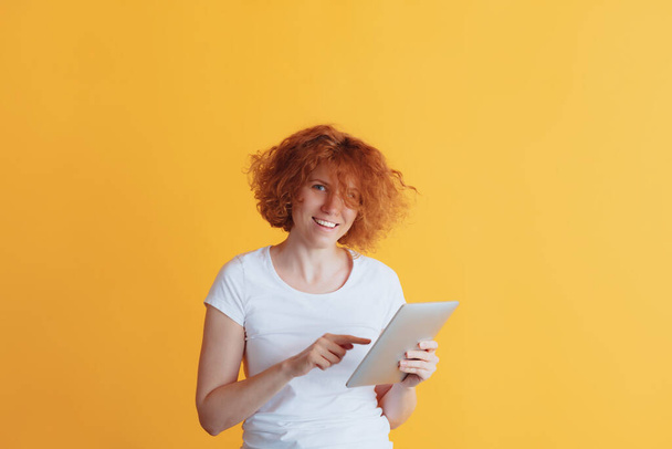 Portrait of smiling woman typing message on tablet or searching information isolated over yellow studio background - Photo, Image