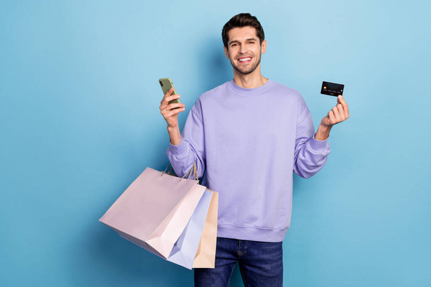 Photo of good young brunet guy hold bags card telephone wear pullover jeans isolated on blue color background - Foto, imagen
