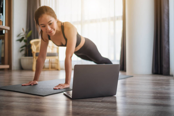Una bella giovane donna asiatica che fa flessioni sul tappeto di formazione mentre guarda tutorial di allenamento online sul computer portatile a casa - Foto, immagini
