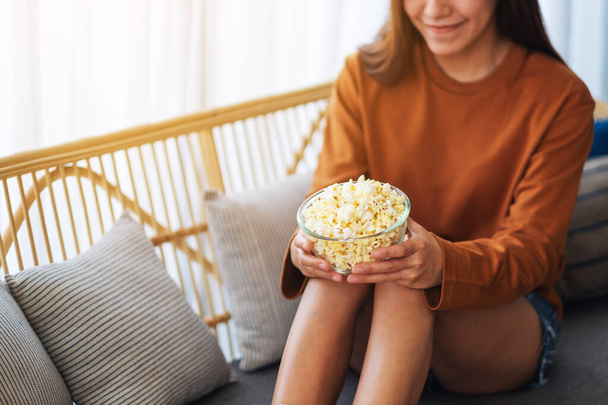 Close up image of a beautiful young woman holding a bowl of popcorn while sitting on sofa at home - Fotó, kép