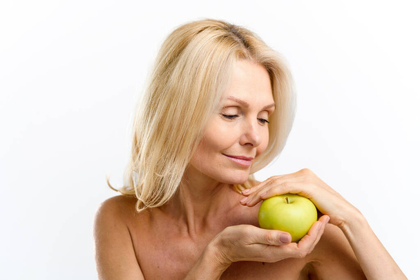 Calm senior woman holding fresh green apple middle her hands and smiling. Elderly model with naked shoulders posing and looking at the fruit isolated on white - Foto, imagen