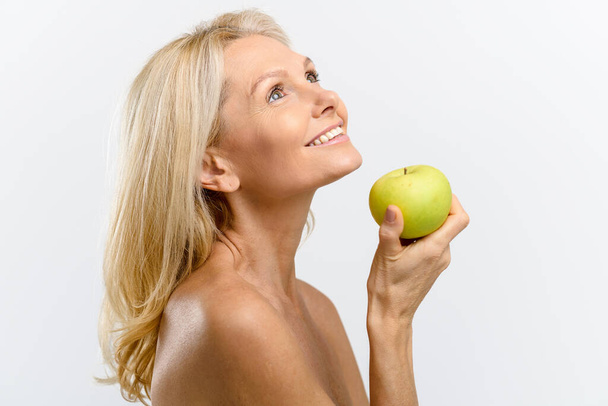 Beautiful and healthy mid age woman with naked shoulders holding green apple near face, looking up and smiling gently - Foto, Imagem