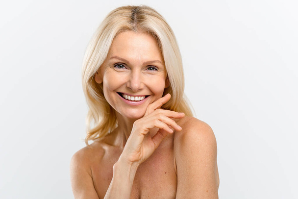 Portrait of fresh and gorgeous middle-aged woman with pure and healthy skin and blonde hair, stands with a naked shoulders isolated on white background, mature lady looks at camera - Foto, Bild