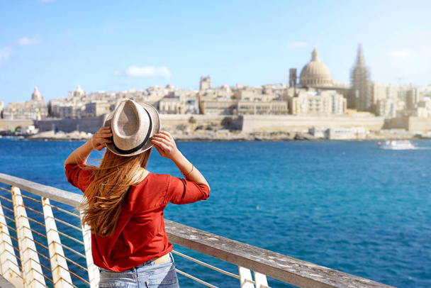 Turismo en Europa. Chica viajera sosteniendo sombrero caminando a lo largo del paseo marítimo de Malta con paisaje urbano de La Valeta en el fondo. - Foto, Imagen