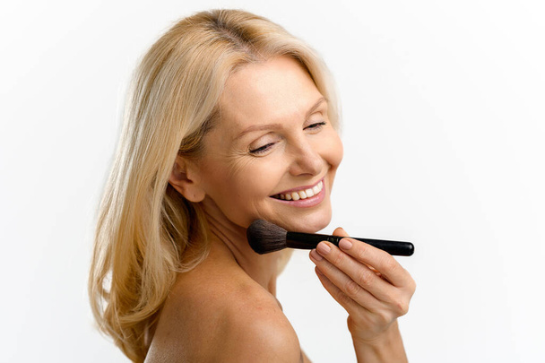 Blonde 40s 50s middle aged woman with holding make up brush on shining face with perfect skin, looking away, applying powder or some cosmetic product - Φωτογραφία, εικόνα