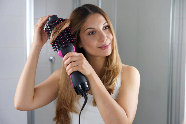 Girl using round brush hair dryer to style hair at the mirror in an easy way at home - Fotó, kép