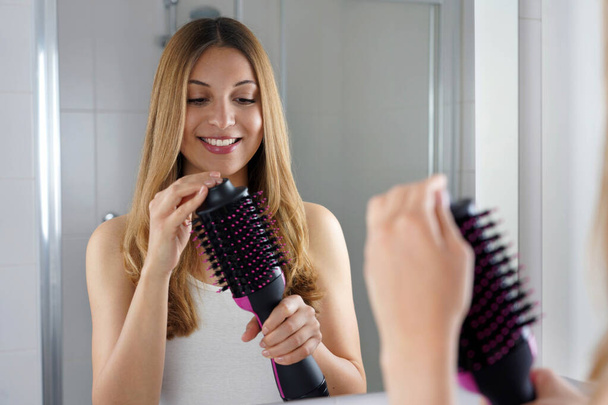 Pleased girl holds round brush hair dryer in her bathroom at home. Young woman holding her salon one-step brush hair dryer and volumizer. - Foto, afbeelding