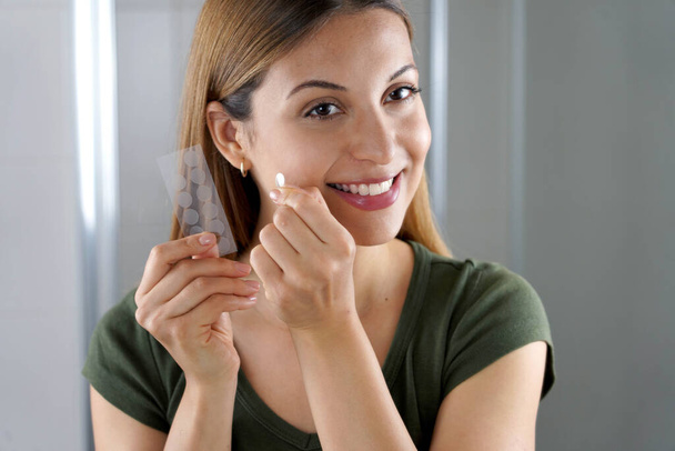 Gros plan de la jeune fille souriante appliquant le patch anti-piquage de traitement de l'acné sur un bouton dans la salle de bain à la maison - Photo, image