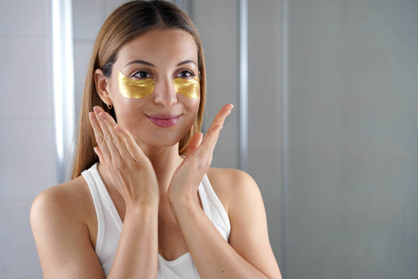 Όμορφο κορίτσι που βάζει χρυσή αντιγηραντική μάσκα κάτω από τα μάτια κοιτάζεται στον καθρέφτη στο μπάνιο. Δερμοκαλλυντική γυναίκα με μπαλώματα υφασμάτινης μάσκας κάτω από τα μάτια για μείωση των ματιών. - Φωτογραφία, εικόνα