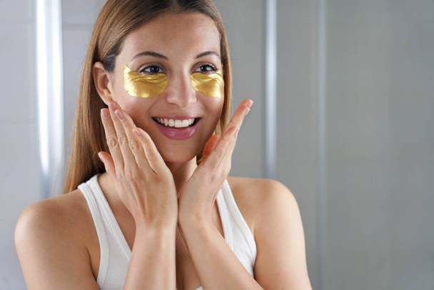Close-up of beauty woman applying golden anti-aging under-eye mask looking herself in the mirror in the bathroom. Skin care girl touch patches of fabric mask under eyes to reduce eye bags. - Fotó, kép