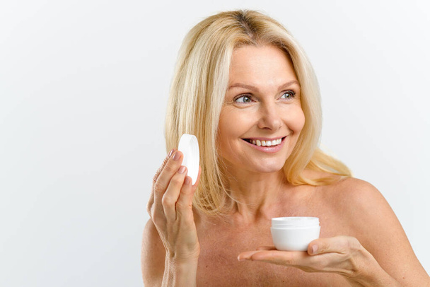 Skincare routine. Middle aged 50s woman holding jar with anti-aging moisturiser, preparing applying cream on the face, looking away isolated on white - Foto, Bild