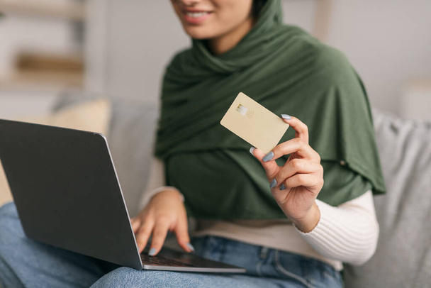 Περικοπή άποψη του χιλιετή αραβική γυναίκα σε μαντίλα χρησιμοποιώντας πιστωτική κάρτα και φορητό υπολογιστή, ψώνια σε απευθείας σύνδεση στον καναπέ στο σπίτι - Φωτογραφία, εικόνα