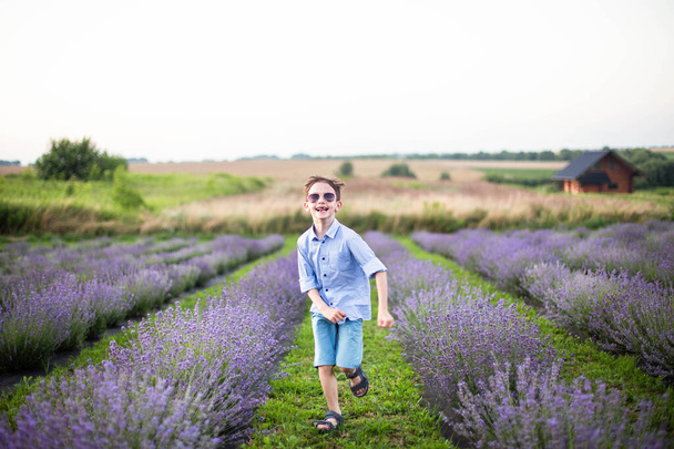 Χαμογελαστό αγοράκι που τρέχει στο χωράφι με τη λεβάντα. Αστείο παιδί με γυαλιά ντυμένο με μπλε πουκάμισο και ψάθινο καπέλο που τρέχει στο γρασίδι ανάμεσα σε λουλούδια λεβάντας. Οριζόντια φωτογραφία - Φωτογραφία, εικόνα