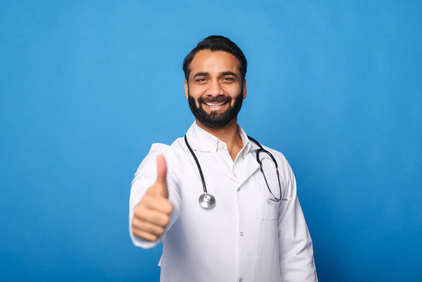 Cheerful Indian male doctor with a stethoscope in a medical gown posing isolated over blue - Фото, изображение