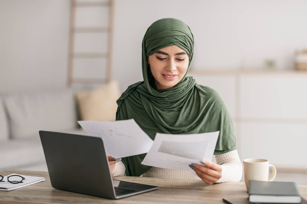 ラップトップを使用してヒジャーブの幸せな若いアラブ人女性は、自宅でオンライン作業会議中にビジネス文書を見て - 写真・画像