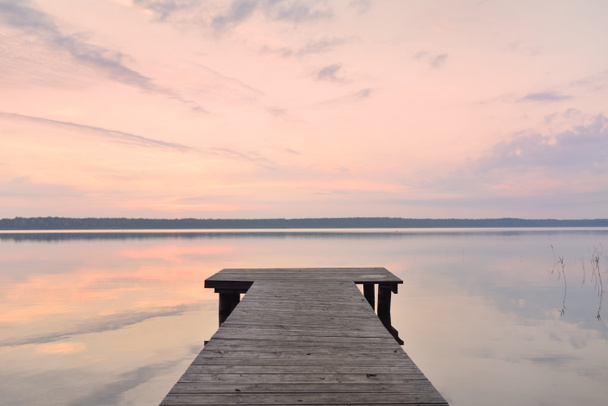 Forest lake at sunset. Wooden pier. Soft sunlight, glowing clouds, symmetry reflections in a crystal clear water. Idyllic landscape. Panoramic view. Nature, ecology, ecotourism. Peace and joy concepts - Photo, image