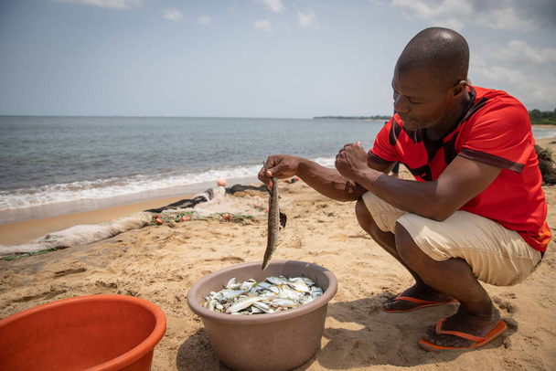 African fisherman looks at the fish he just caught in a bucket, fisher at the beach while working - Zdjęcie, obraz