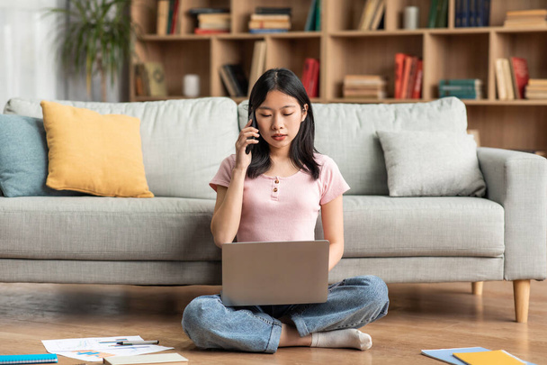 Asian female freelancer talking on cellphone and typing on laptop, working online from home, sitting on floor - Photo, Image