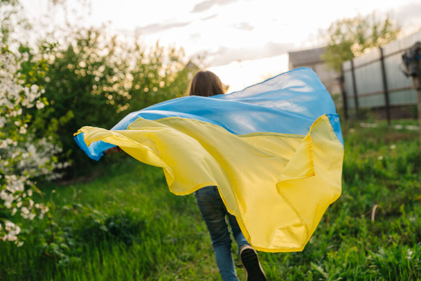 A young girl runs with the Ukrainian flag. Photo of a girl running with a Ukrainian flag used as a cape - Photo, Image