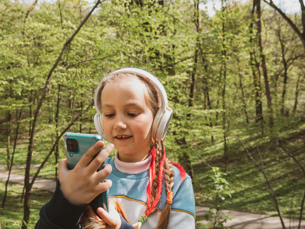 Adorable child girl in headphones learning language listening to music audio books podcasts smartphone outdoor. Blogger kid with gadget distance school online lesson chat cell phone conference in park - Foto, afbeelding