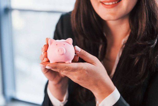 Woman in black formal clothes is indoors in the modern office holds pink piggy bank. - Φωτογραφία, εικόνα