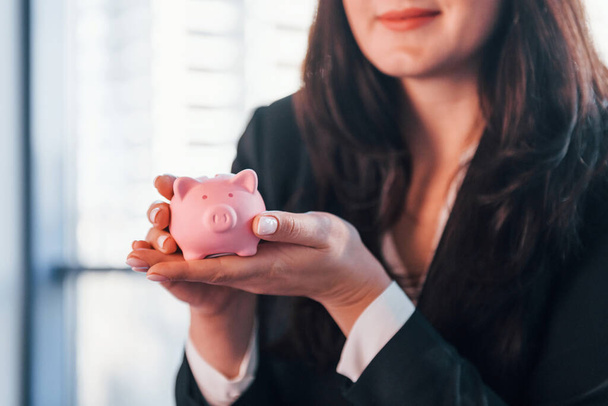 Woman in black formal clothes is indoors in the modern office holds pink piggy bank. - Foto, immagini